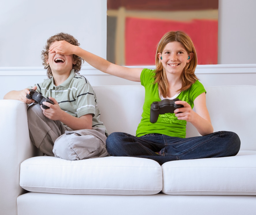 A girl with hearing aids and bright green t-shirt and a boy sitting on a sofa, each holding a gaming pad, engaged in a game.